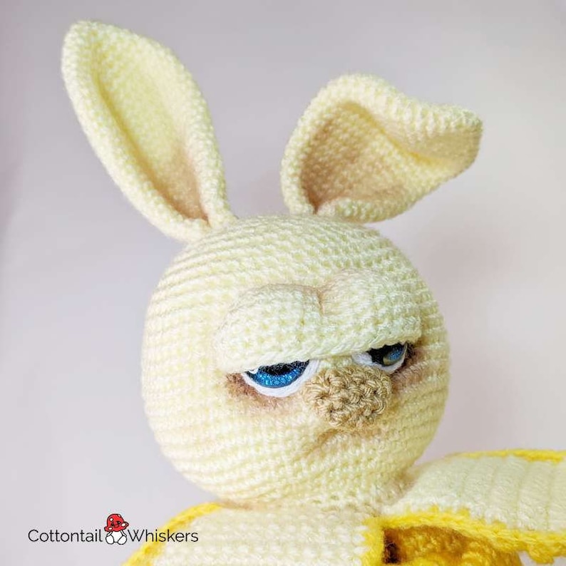 May include: A crocheted yellow bunny with a grumpy expression. The bunny has blue eyes and a brown nose. The bunny is wearing a yellow crocheted outfit.