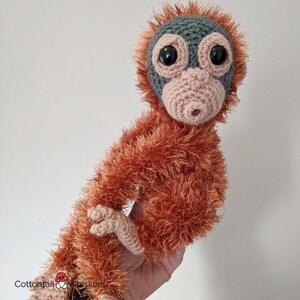 May include: A crocheted orangutan plush toy with a light brown fur, a pink nose, and large black eyes. The toy is sitting on a hand.