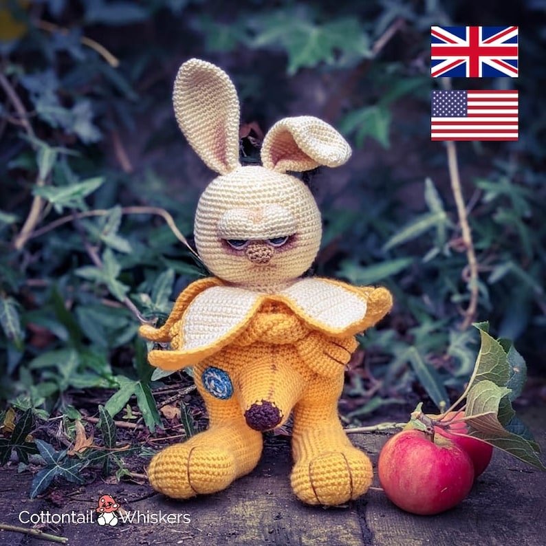 May include: A crocheted banana-shaped bunny with a grumpy expression. The bunny is wearing a yellow banana costume with a brown stem. The bunny is sitting on a wooden surface with two red apples.