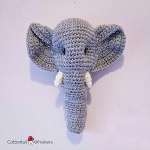 Peut inclure: Un jouet en forme d'éléphant en crochet gris avec des défenses en crochet blanc. L'éléphant est accroché à un mur.