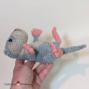 May include: A crocheted gray and pink otter plush toy with a long tail. The toy is lying on its back with its legs and feet in the air.
