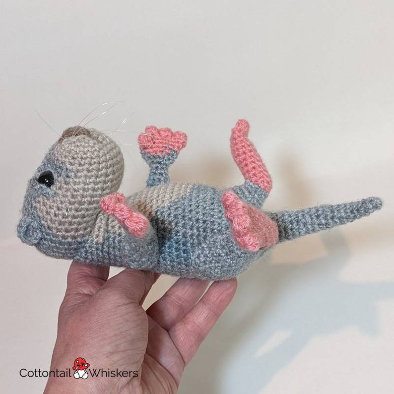 May include: A crocheted gray and pink otter plush toy with a long tail. The toy is lying on its back with its legs and feet in the air.