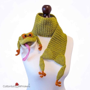 May include: A handmade green crochet scarf shaped like a frog, displayed on a mannequin. The scarf features a frog head with orange eyes and a pink tongue, and frog legs. The scarf is approximately 60 inches long.