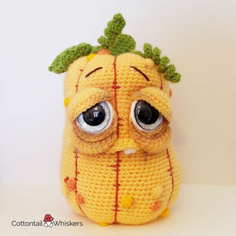 May include: A crocheted plush toy shaped like a pumpkin with a sad expression. The pumpkin is yellow with orange stitching and has green leaves on top. The toy has large, black eyes with silver glitter.