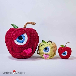 Op de afbeelding: Drie gehaakte appelknuffels, één rood, één groen en één kleinere rood. Elke appel heeft één groot oog en houdt een klein roze hart vast.