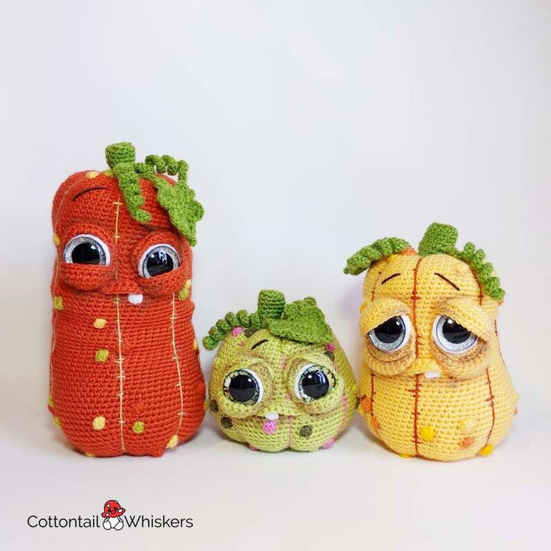 May include: Three crocheted pumpkins with different facial expressions. The largest pumpkin is orange with yellow and red accents, the middle pumpkin is green with pink and brown accents, and the smallest pumpkin is yellow with orange accents.