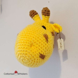 Puede incluir: Una cabeza de jirafa de crochet colgada en la pared con manchas marrones y una etiqueta de madera que dice "Glenda".