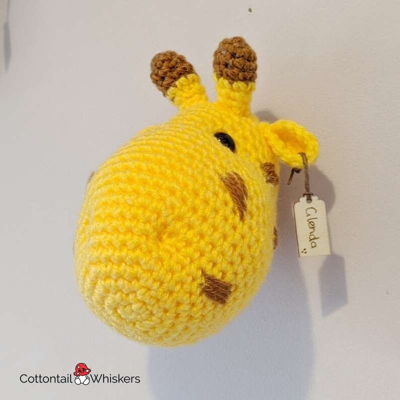 Puede incluir: Una cabeza de jirafa de crochet colgada en la pared con manchas marrones y una etiqueta de madera que dice "Glenda".