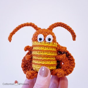 May include: A small, handcrafted crochet insect toy featuring a rust-orange body with two long antennae, white eyes with black pupils, and a central abdomen section with alternating horizontal stripes of bright yellow and rust-orange yarn.