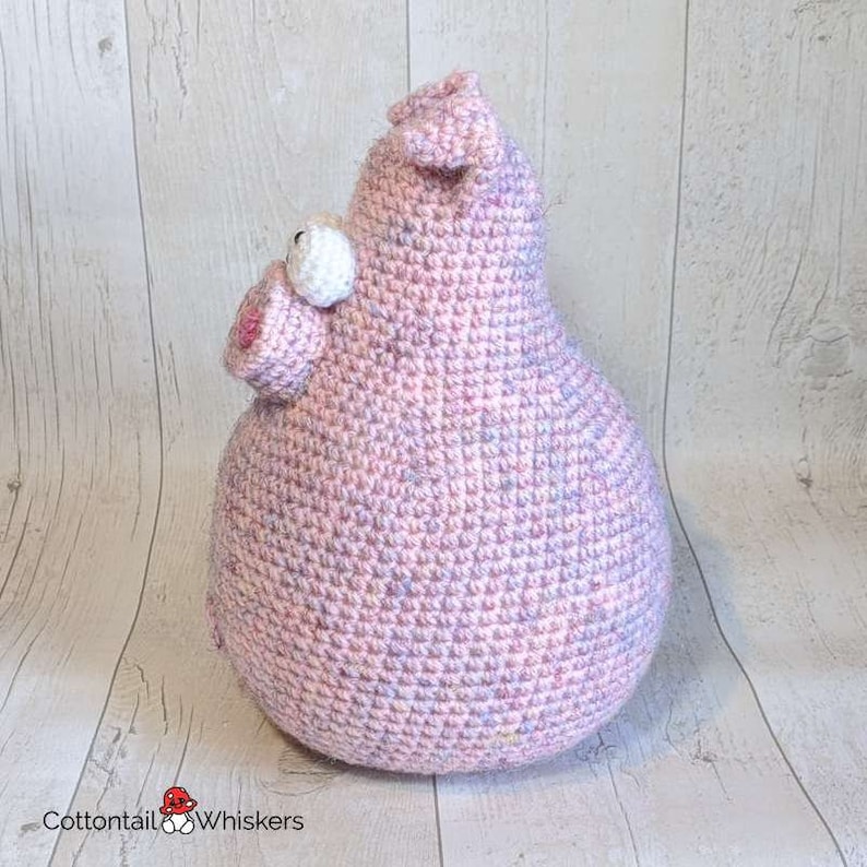 Op de afbeelding: Een roze gehaakt knuffelvarken met een roze neus en witte ogen. Het varken zit op een wit houten oppervlak.