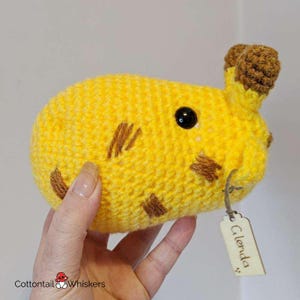 Puede incluir: Un animal de peluche de crochet amarillo con detalles marrones y un ojo negro. El animal tiene una etiqueta que dice "Glenda".