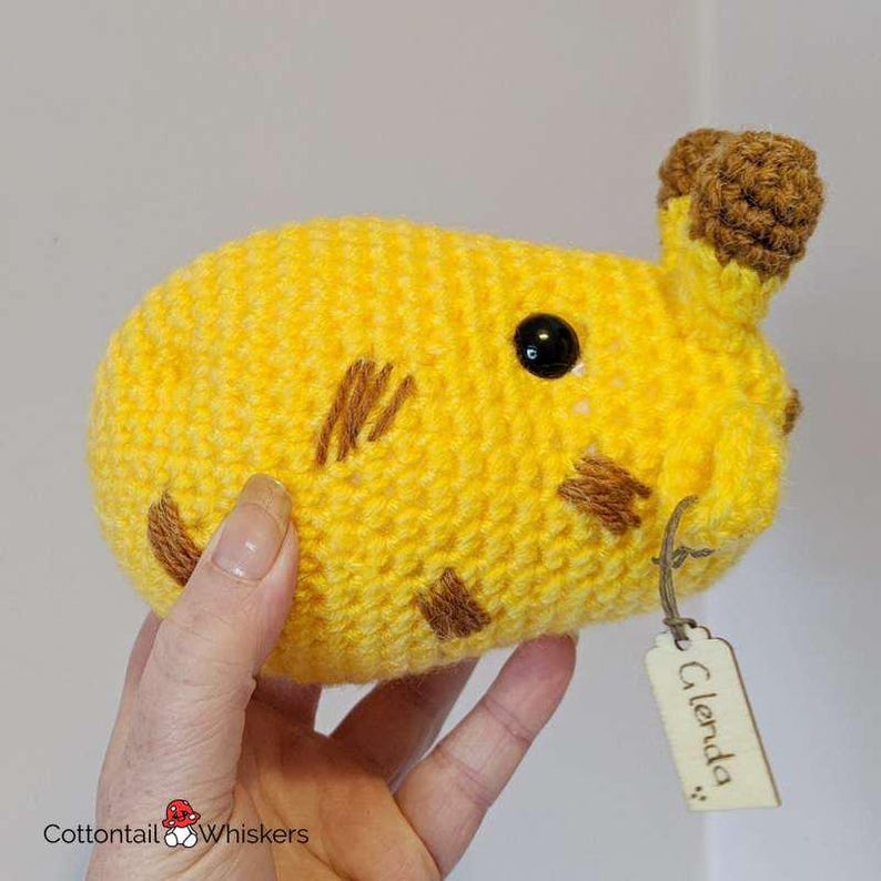 Puede incluir: Un animal de peluche de crochet amarillo con detalles marrones y un ojo negro. El animal tiene una etiqueta que dice "Glenda".