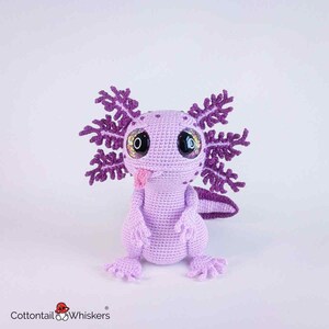 May include: A handmade crochet axolotl in shades of lavender and purple. The plush toy features large, glittery eyes, a pink tongue, and detailed gills. The axolotl is a whimsical, soft sculpture, perfect for a child's room or as a unique gift.