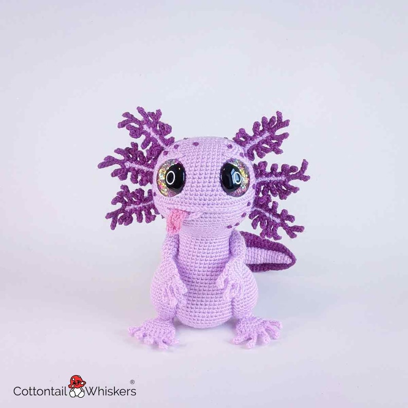 May include: A handmade crochet axolotl in shades of lavender and purple. The plush toy features large, glittery eyes, a pink tongue, and detailed gills. The axolotl is a whimsical, soft sculpture, perfect for a child's room or as a unique gift.