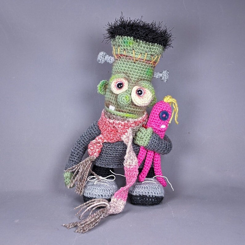 Pode incluir: Um boneco de Frankenstein de croch&ecirc; verde vestindo um su&eacute;ter cinza e uma echarpe rosa e marrom. O monstro est&aacute; segurando um boneco de polvo de croch&ecirc; rosa com um olho de bot&atilde;o azul.