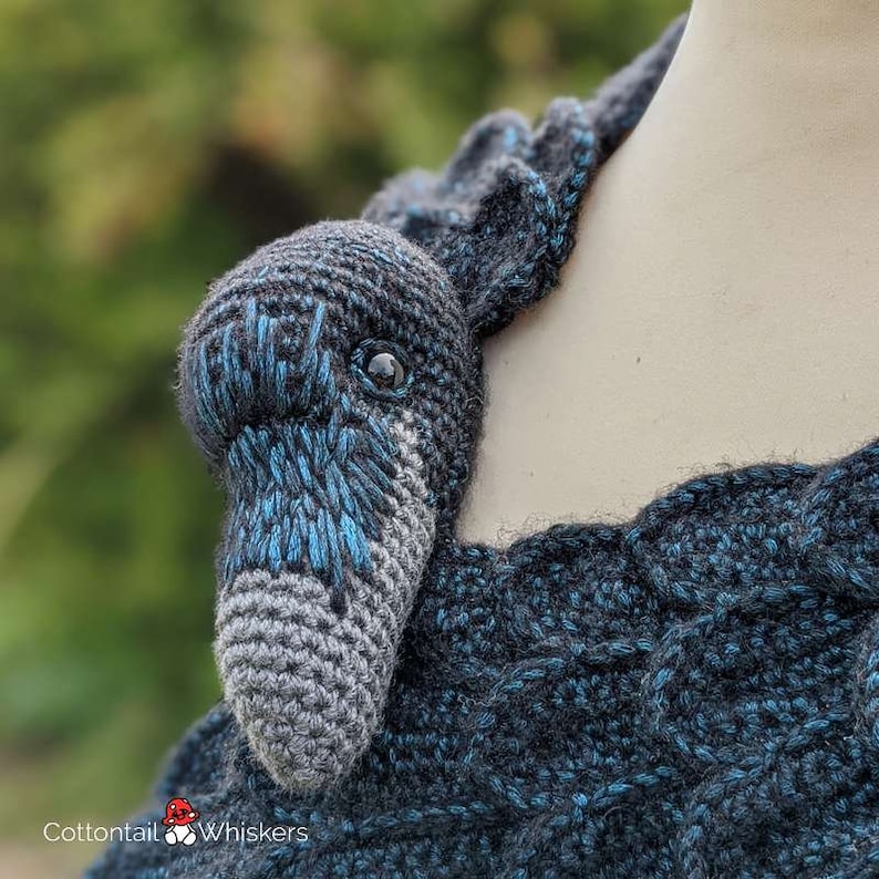 May include: A crocheted raven head with blue and gray yarn, attached to a black and blue crocheted scarf. The scarf is draped over a white mannequin.