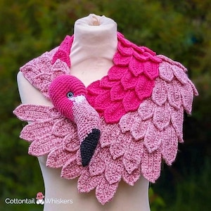 May include: A pink crocheted flamingo scarf with a 3D flamingo head attached. The scarf is made of pink yarn and has a scalloped edge. The flamingo head is pink with black eyes and a black beak.