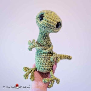 May include: A handmade crochet lizard in shades of green and yellow. The lizard has large black eyes and is posed with its arms crossed. The lizard's feet and tail are also visible. The logo "Cottontail & Whiskers" is in the bottom left corner.