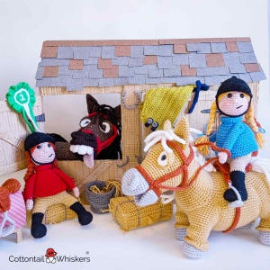 May include: A crocheted horse and two crocheted dolls in riding outfits. The horse is brown and white with a red bridle. The dolls are wearing red and blue riding outfits with helmets. The dolls are standing in front of a wooden stable with a hay bale and a bucket.