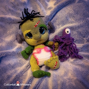 May include: A handmade crochet Frankenstein's monster doll with a purple octopus toy. The doll has green skin, black hair, and a pink and white diaper. The octopus is purple with one large eye. The toys are on a blue blanket.