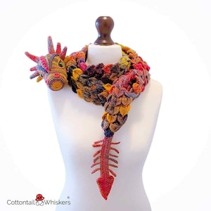Op de afbeelding: Een kleurrijke, gehaakte draken sjaal, tentoongesteld op een mannequin. De sjaal heeft een drakenkop met hoorns en een staart. De sjaal is gemaakt van verschillende kleuren, waaronder rood, geel, blauw en paars. Het logo "Cottontail & Whiskers" staat linksonder.