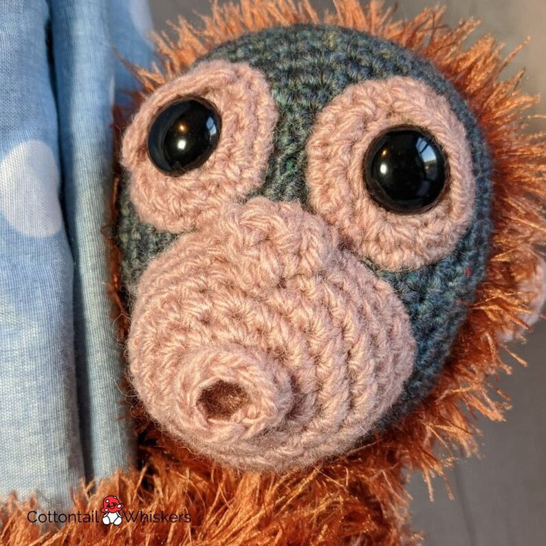 May include: Close-up of a crocheted orangutan plush toy with brown fur, large black eyes, and a pink crocheted nose. The toy is partially visible against a blue and white patterned background.