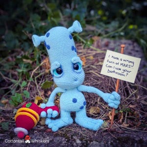 May include: A light blue crocheted alien doll with dark blue spots and large eyes. The alien holds a red and yellow crocheted object and a sign that reads, "I took a wrong turn at MARS... Can I use your bathroom?"