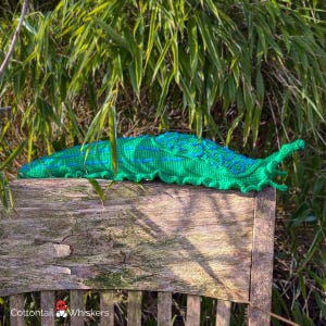 K&ouml;nnte beinhalten: Ein gr&uuml;ner geh&auml;kelter Schleim mit blauen Akzenten sitzt auf einem Holzstuhl. Der Schleim liegt auf der Seite, Kopf und Schwanz sind sichtbar. Der Stuhl ist aus verwittertem Holz und hat eine Lattenlehne.