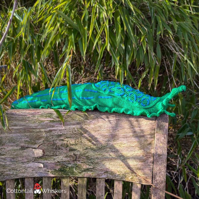 K&ouml;nnte beinhalten: Ein gr&uuml;ner geh&auml;kelter Schleim mit blauen Akzenten sitzt auf einem Holzstuhl. Der Schleim liegt auf der Seite, Kopf und Schwanz sind sichtbar. Der Stuhl ist aus verwittertem Holz und hat eine Lattenlehne.