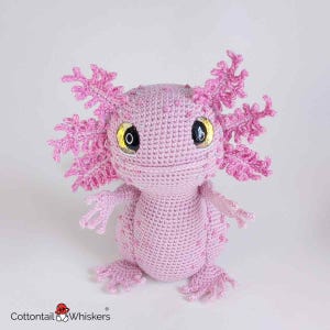 May include: A handmade pink crochet axolotl plush toy with detailed gills and limbs. The axolotl has large, expressive eyes with black pupils and yellow irises. The plush is made of soft yarn and is a cute, whimsical creature.