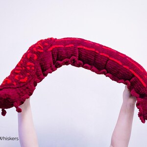K&ouml;nnte beinhalten: Ein gro&szlig;er, geh&auml;kelter, roter Wurm mit einer strukturierten Oberfl&auml;che. Der Wurm wird von zwei H&auml;nden hochgehalten.