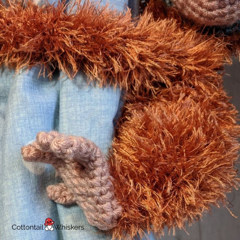 May include: Close-up of a crocheted orangutan arm with brown fur. The arm is bent at the elbow and the hand is open. The fur is very fluffy and soft. The hand is crocheted in a light brown color.