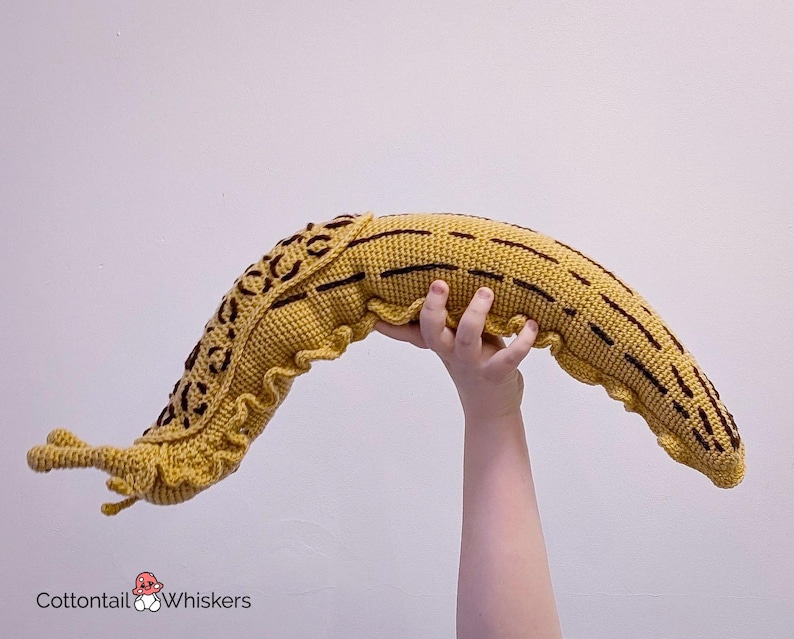 K&ouml;nnte beinhalten: Ein geh&auml;keltes Pl&uuml;schtier in Form einer Schnecke in Gelb mit braunen Streifen. Die Schnecke wird von einer Hand hochgehalten.