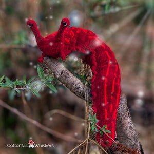 K&ouml;nnte beinhalten: Ein roter geh&auml;kelter Wurm mit einer strukturierten Oberfl&auml;che, der auf einem Ast mit gr&uuml;nen Bl&auml;ttern sitzt. Der Wurm hat einen langen, schlanken K&ouml;rper und einen spitzen Kopf. Der Hintergrund ist verschwommen und unscharf.