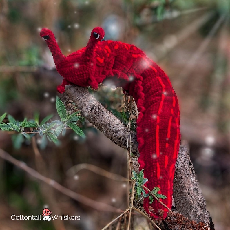 K&ouml;nnte beinhalten: Ein roter geh&auml;kelter Wurm mit einer strukturierten Oberfl&auml;che, der auf einem Ast mit gr&uuml;nen Bl&auml;ttern sitzt. Der Wurm hat einen langen, schlanken K&ouml;rper und einen spitzen Kopf. Der Hintergrund ist verschwommen und unscharf.