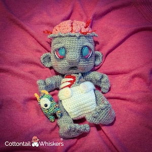 May include: A handmade crochet zombie doll with a pink brain, blue eyes, and a white diaper. The doll is gray with red accents and holds a small, colorful crochet monster. The doll is on a pink background. The text "Cottontail Whiskers" is in the bottom left corner.