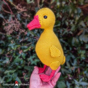 May include: A handmade crochet duck toy featuring a bright yellow body, a textured orange beak, and matching orange webbed feet. The duck has small black bead eyes and is crafted with a visible yarn stitch pattern throughout its rounded form.