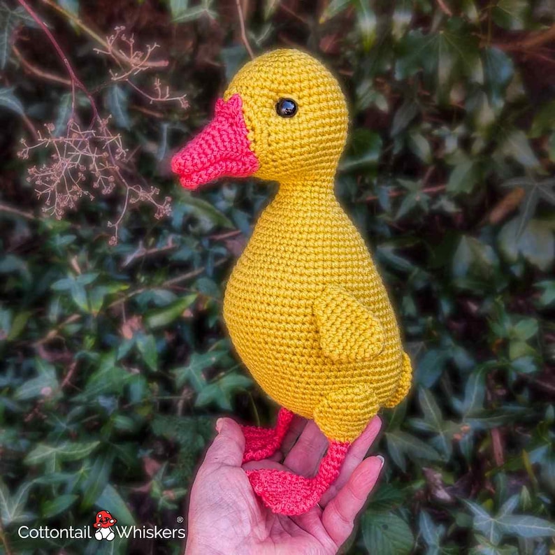 May include: A handmade crochet duck toy featuring a bright yellow body, a textured orange beak, and matching orange webbed feet. The duck has small black bead eyes and is crafted with a visible yarn stitch pattern throughout its rounded form.