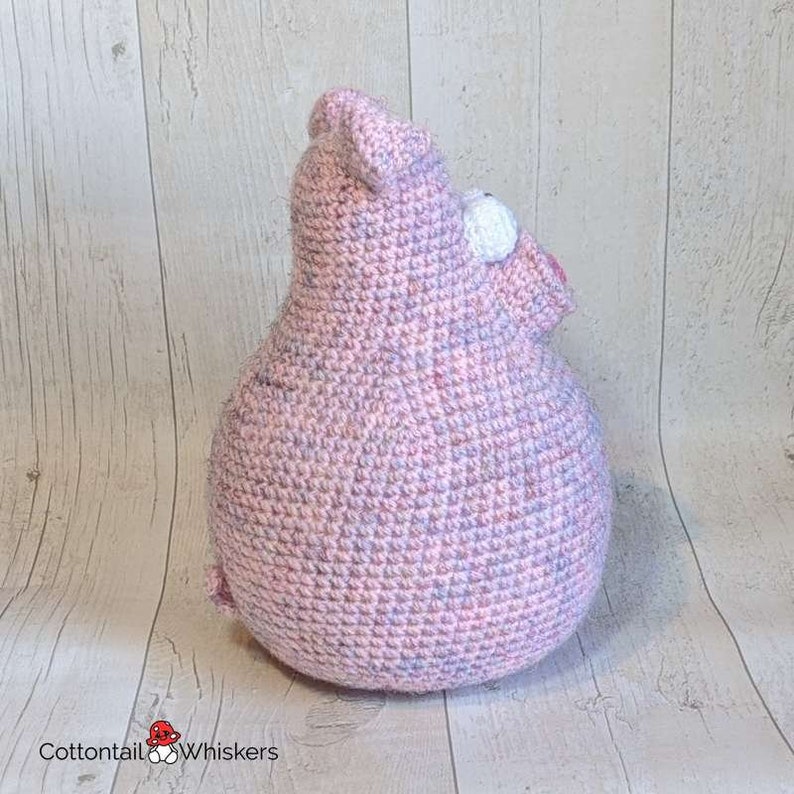 Op de afbeelding: Een roze en paarse gehaakte varkens knuffel. Het varken zit op een licht houten achtergrond.
