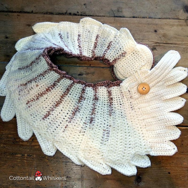 Pu&ograve; includere: Un collo in crochet bianco con strisce marroni e un bottone in crochet marrone. Il collo &egrave; steso piatto su una superficie di legno.