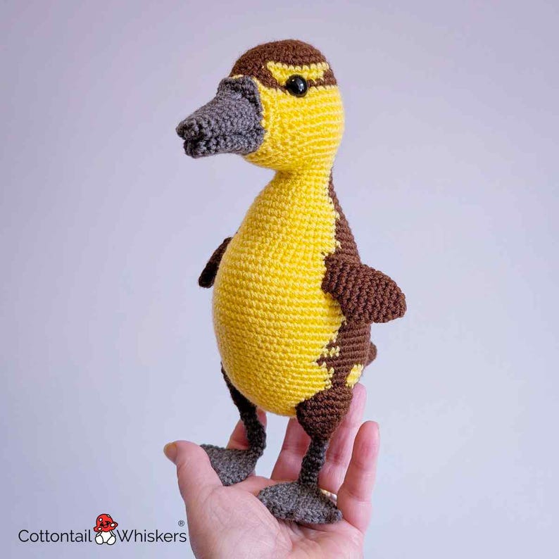May include: A handmade crochet duckling toy in yellow and brown. The duckling has a yellow body with brown accents on the wings and back. It has a gray beak and feet, and black eyes. The toy is held in a hand.