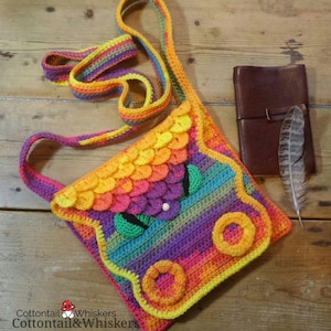 May include: A rainbow crochet crossbody bag with a dragon face design. The bag has a yellow, orange, red, purple, blue, and green color scheme. The dragon face has green eyes and a white nose.