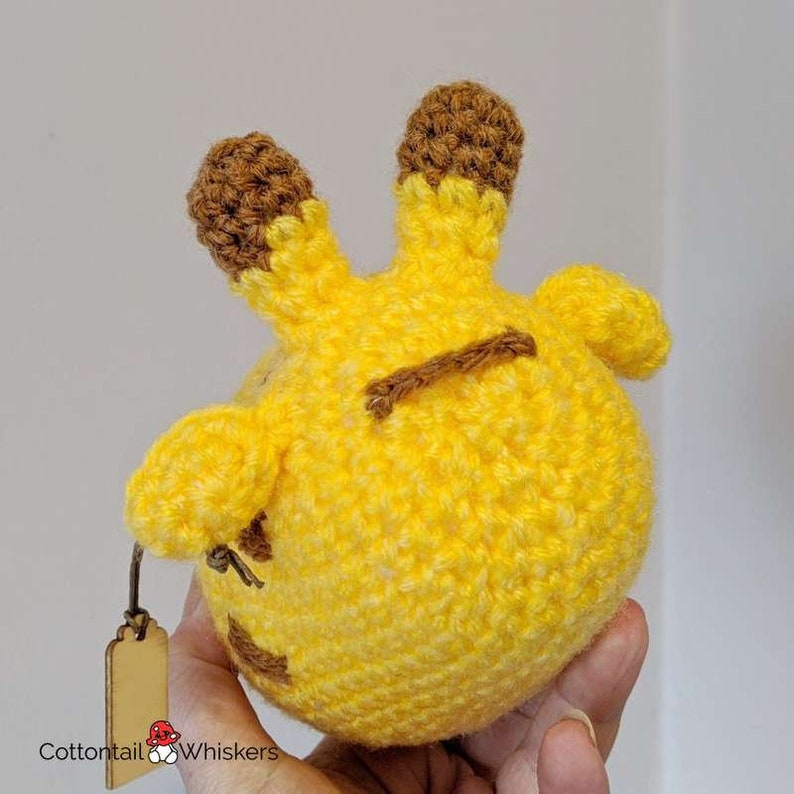 Puede incluir: Un animal de peluche de crochet con forma de jirafa. La jirafa es amarilla con orejas marrones y una franja marr&oacute;n en la espalda. La jirafa est&aacute; siendo sostenida en una mano.