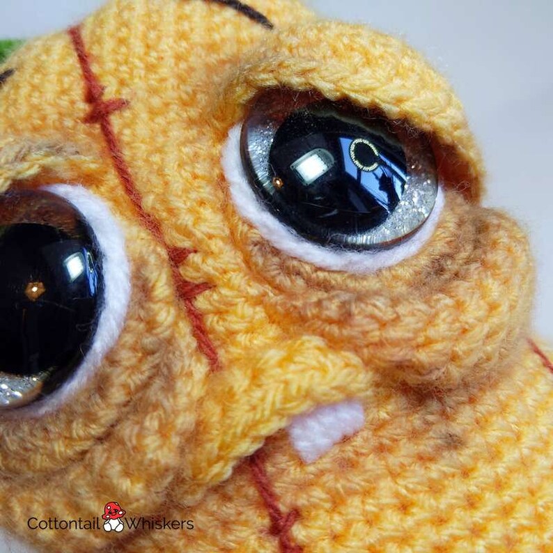 May include: Close-up of a crocheted orange and brown plush toy with large black eyes and a white stitched mouth. The toy has red stitching details on the face.