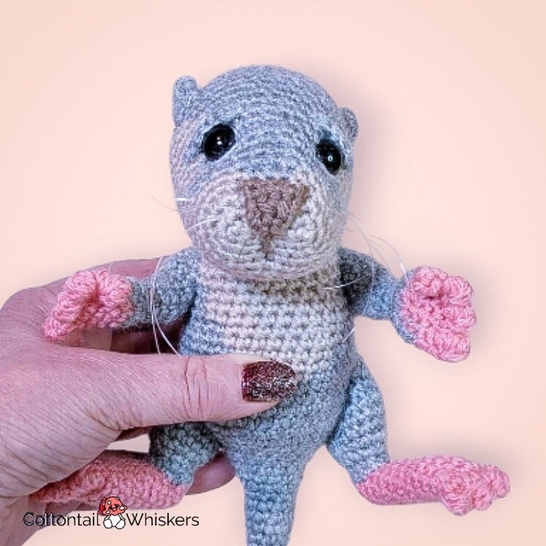 May include: A crocheted gray and pink otter plush toy with black eyes and a brown nose. The toy is being held by a hand with pink nail polish.