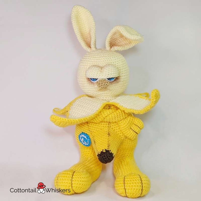 May include: A crocheted stuffed toy shaped like a banana with a bunny head. The bunny has blue eyes and a yellow body. The banana is yellow with a brown stem.