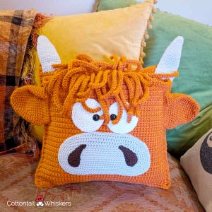 May include: A square crochet pillow shaped like a Highland cow with orange yarn, white horns, and a white muzzle. The cow features long, shaggy orange hair, two black button eyes, and dark brown nostrils. The text Cottontail Whiskers is visible.