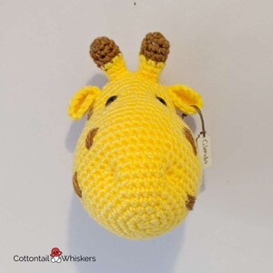 Puede incluir: Una cabeza de jirafa de crochet para colgar en la pared con orejas y manchas marrones. La jirafa es amarilla y tiene una etiqueta que dice "Glenda".