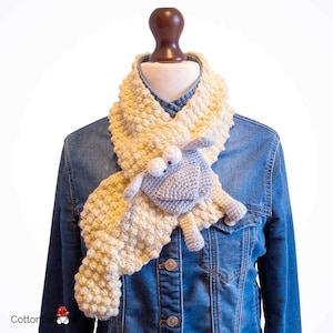 May include: A handmade, cream-colored crochet scarf with a textured, bobble stitch design. The scarf features a gray crochet sheep applique with white eyes. The scarf is displayed on a mannequin wearing a denim jacket.