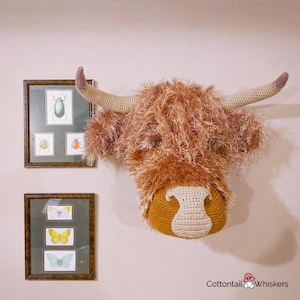 Op de afbeelding: Een gehaakte Highland Cow-kop met een pluche bruine en witte vachttextuur. De koppen worden aan de muur gemonteerd met twee ingelijste afbeeldingen van insecten en vlinders.